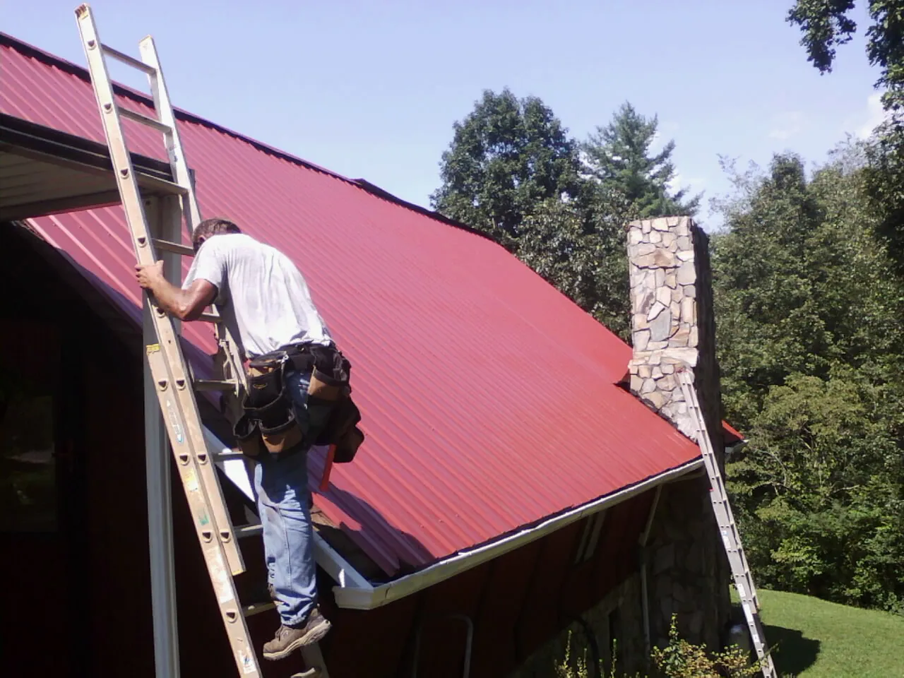 Roofer on a ladder beside a red metal roof and stone chimney — Soffit & Fascia Repair by Roofing Brothers in Lowpoint